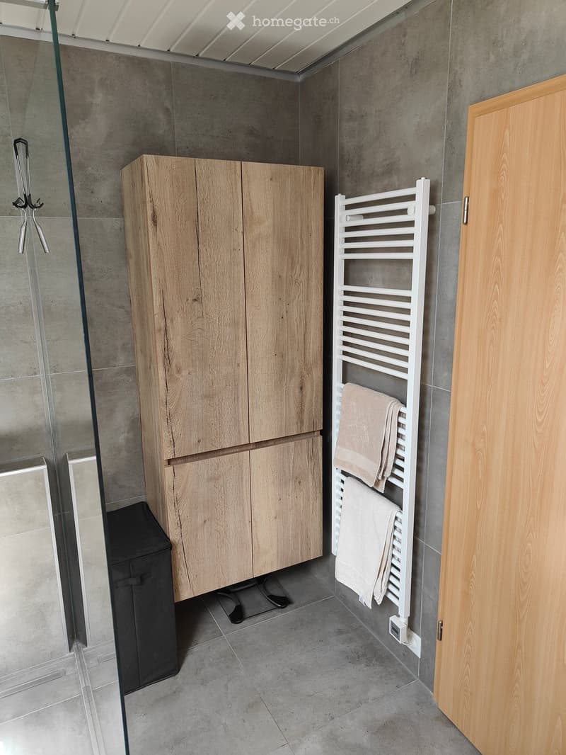 Modern bathroom with wooden cabinet, white towel rack with towels, grey tiled walls, and a glass shower enclosure.