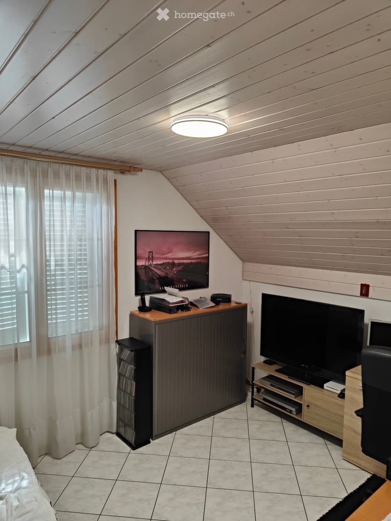 A cozy attic room with a slanted wooden ceiling, a bed, a large window with white curtains, a TV on a stand, and a wooden cabinet with a printer and other items.