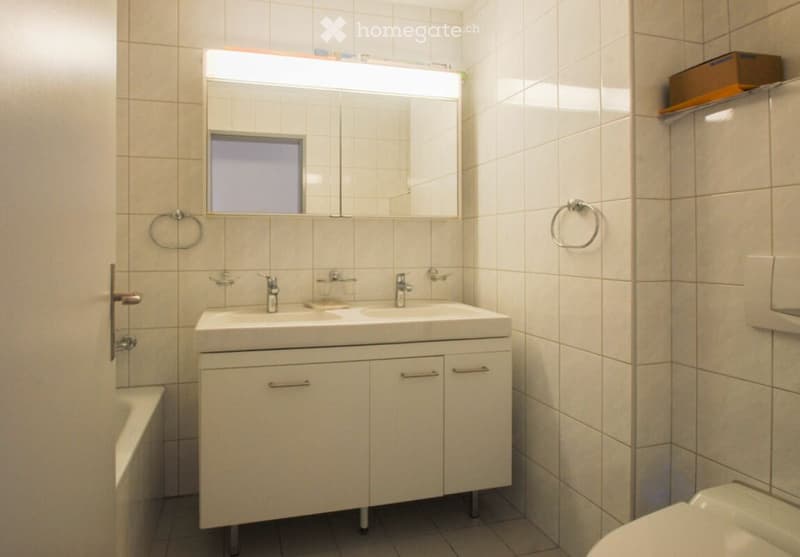 Modern bathroom with a double sink, large mirror, white tiled walls, and a toilet. Features include a towel ring, cabinet storage, and a frosted window above the sink.