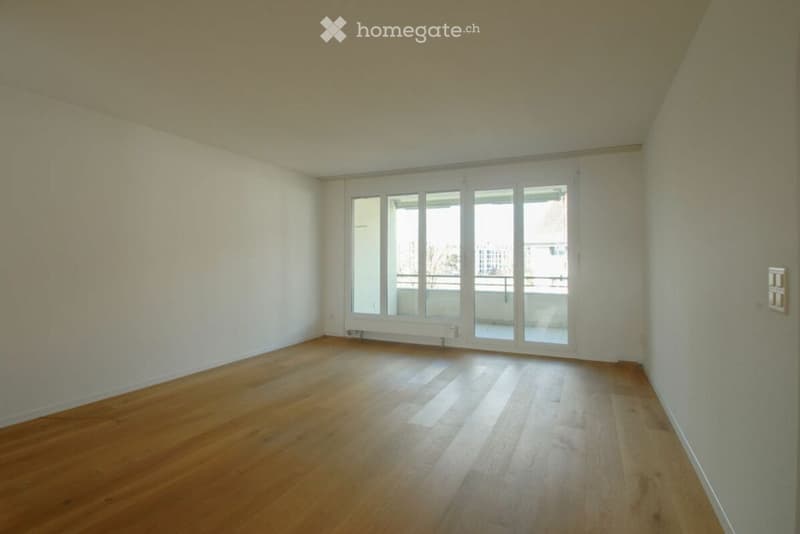 Empty room with wooden flooring, white walls, large windows, and a sliding glass door leading to a balcony.