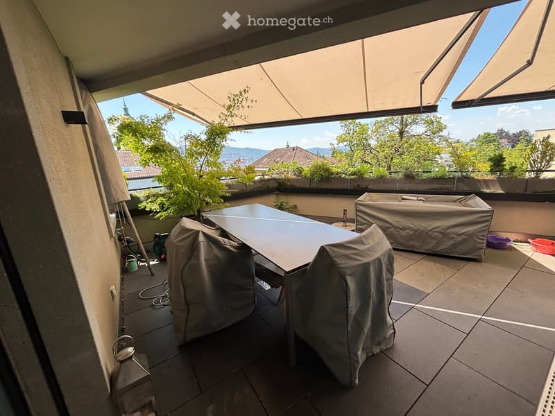 Balcony with a covered table and chairs, overlooking a cityscape with trees and mountains in the background.