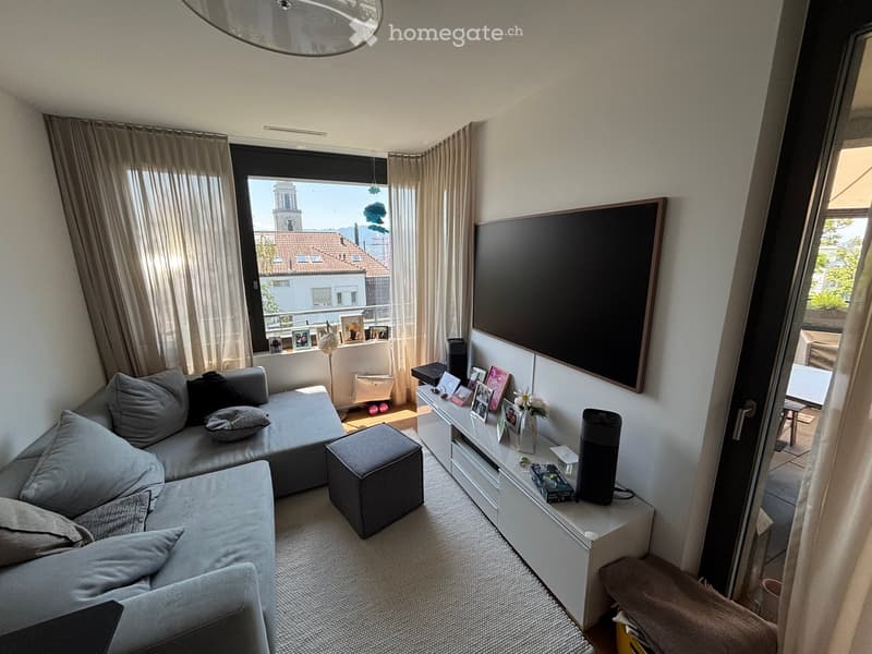 Modern living room with a grey sectional sofa, ottoman, white side table, television, and large windows with curtains. The room features a view of a cityscape with a church tower.