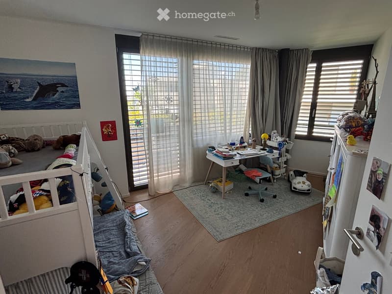 A well-lit bedroom with a lofted bed, desk, chair, and various toys and decorations. The room features large windows with curtains, a wooden floor, and a mix of furniture and personal items.