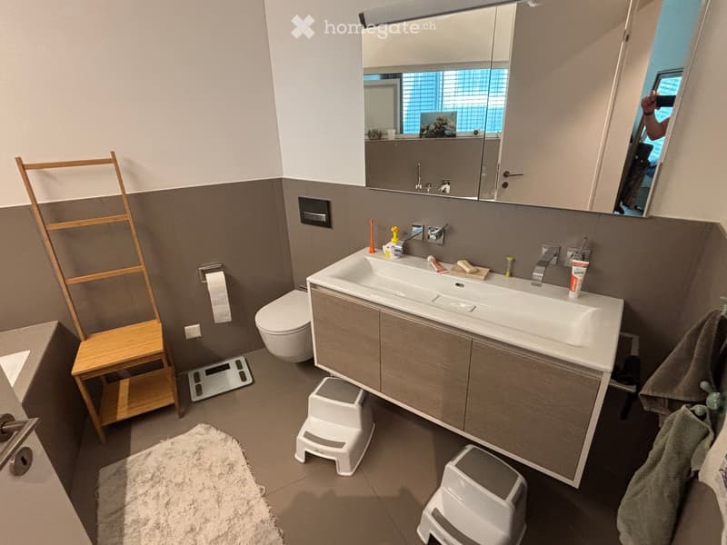 Modern bathroom with a large sink, mirror cabinet, toilet, and wooden chair. Includes a scale, towel, and small stools.