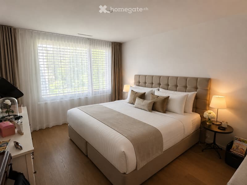 Elegant bedroom with a large bed, tufted headboard, multiple pillows, and a nightstand with a lamp. A side table with a flower vase and a mirror is visible, along with a curtained window and a small table with a lamp.