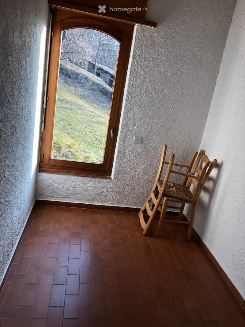 A small, narrow room with terracotta tiled flooring, white textured walls, and a wooden-framed window with a view of a green landscape. A single wooden chair with a woven seat is positioned near the window.