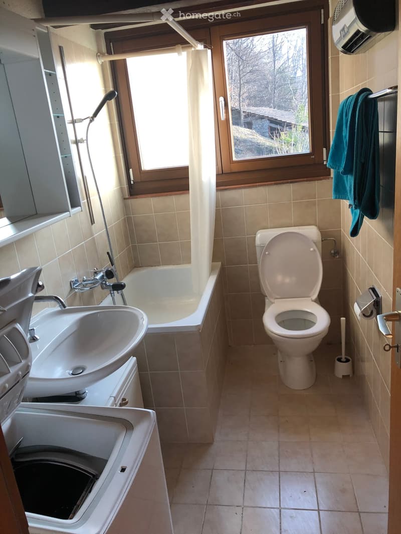Bathroom with beige tiles, a bathtub, a toilet, a sink, a showerhead, and a towel hanging on a rack. A window with a view of a natural setting is visible.