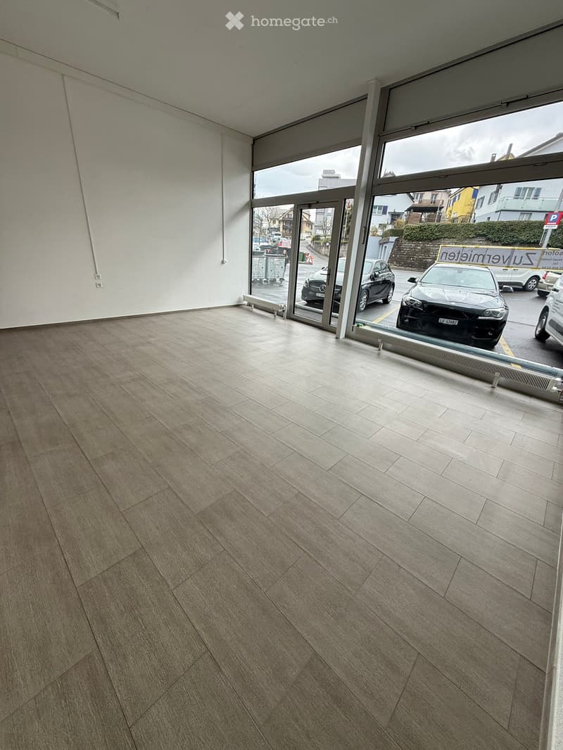 Empty room with light-colored tiled flooring and large glass windows showing a parking area outside.