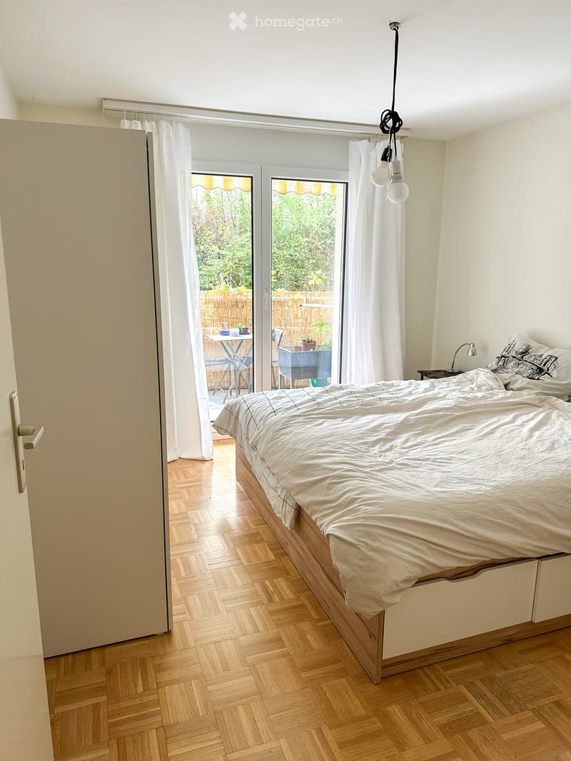 Bright bedroom with wooden flooring, large bed with white bedding, nightstand with lamp, and large windows with white curtains leading to a patio area with outdoor furniture.