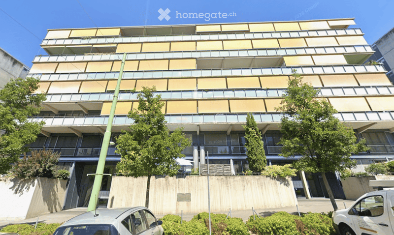 Modern multi-story building with balconies and large windows, surrounded by greenery and parked cars.