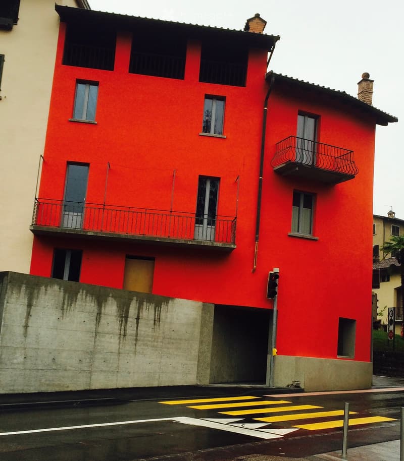 A three-story red building with multiple windows and balconies, located on a street with a pedestrian crossing.