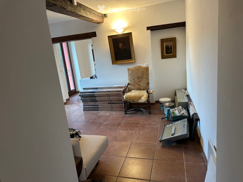 A furnished room with a sofa, a patterned chair, a small table, and a stack of boxes. The room has tiled flooring and white walls with framed paintings.