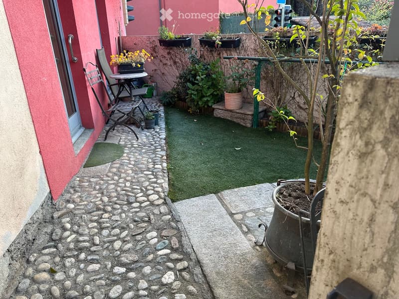 Small garden area with artificial grass, potted plants, a small table, and chairs, surrounded by a stone pathway and a pink building wall.