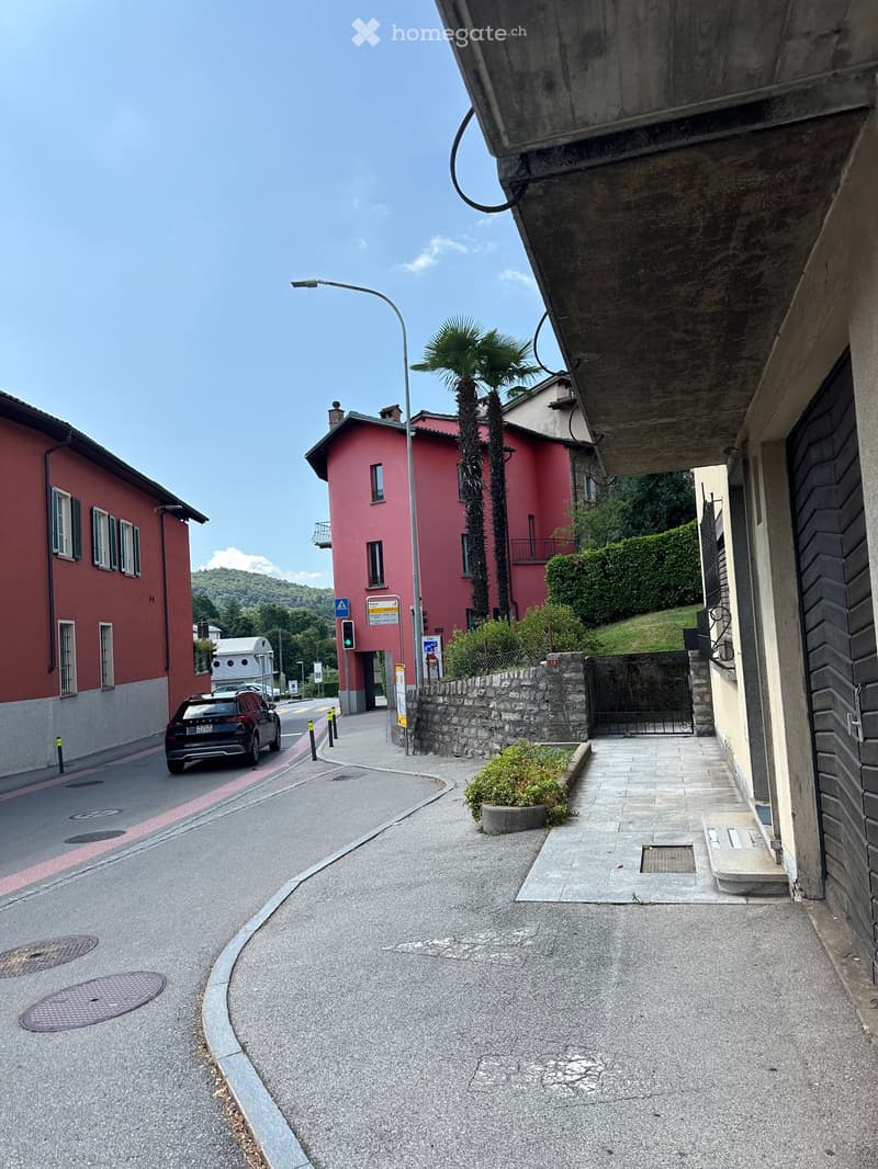 A narrow street lined with colorful buildings, including a pink house with balconies and a red brick building. A palm tree is visible in front of the pink house, and a car is parked on the left side of the road.