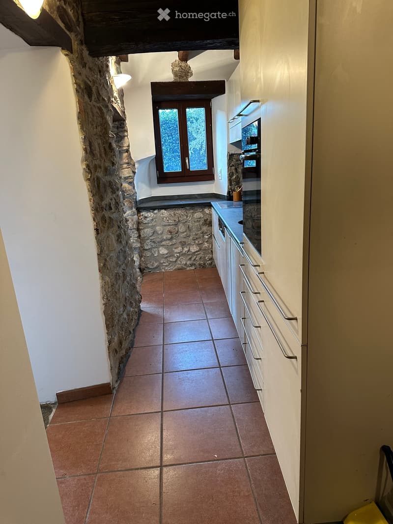 A narrow hallway with tiled flooring, white walls, and a stone wall on one side. A window with wooden frames is visible at the end of the hallway, and a small kitchen area is partially visible on the right side.