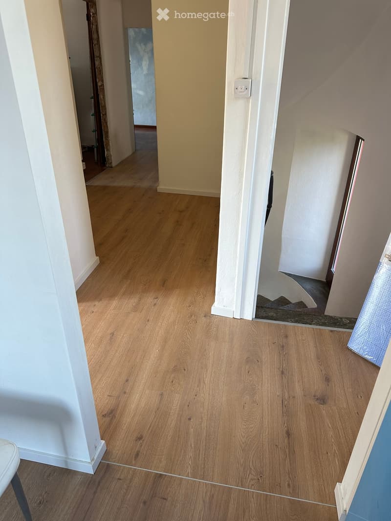 Empty hallway with light wooden flooring and white walls, featuring a visible staircase in the background.