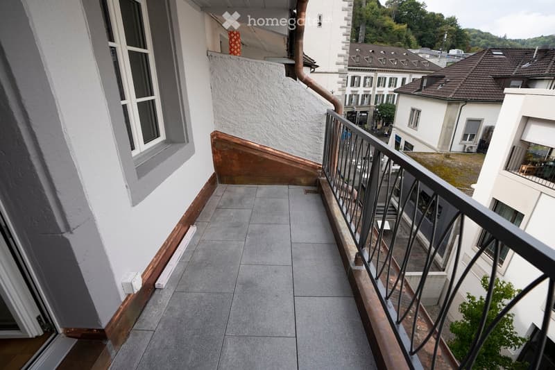 Balcony with grey tiled floor, metal railing, and view of neighboring buildings and greenery.