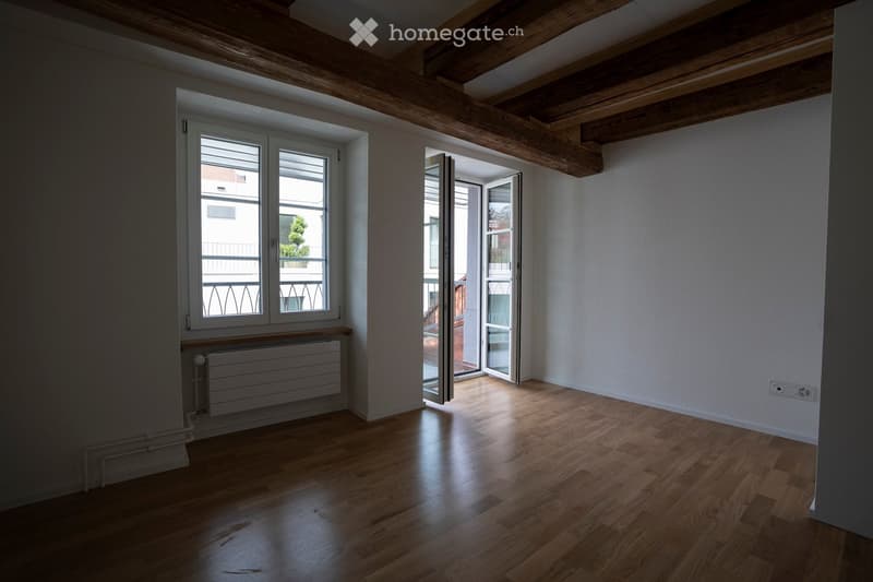 Empty room with wooden flooring, exposed wooden beams on the ceiling, large windows, and a radiator. The room has a balcony access through a glass door.