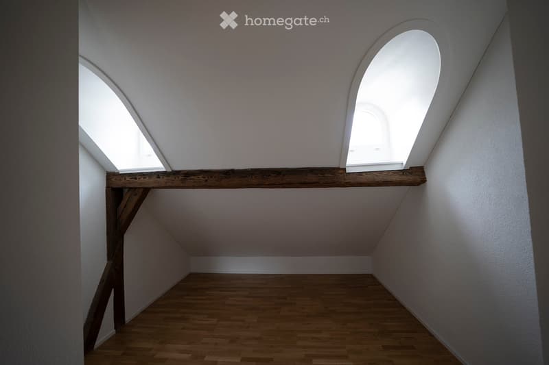 An empty attic space with wooden flooring, white walls, and skylight windows allowing natural light to enter. The room features exposed wooden beams and a simple, minimalist design.