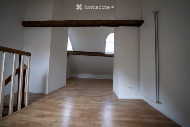 Empty room with wooden flooring, white walls, and a wooden railing leading to an attic area. The attic has exposed wooden beams and small arched windows.