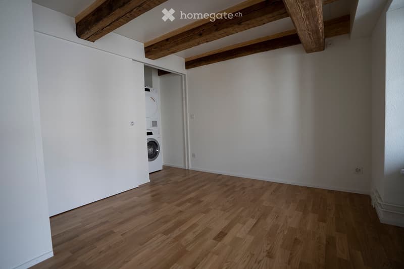 Empty room with wooden flooring, white walls, and exposed wooden beams on the ceiling. A small closet contains a washing machine and dryer.