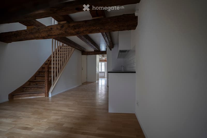Bright hallway with wooden flooring, exposed wooden beams, and a staircase leading upwards. Minimalist design with white walls and a small white cabinet on the right side.