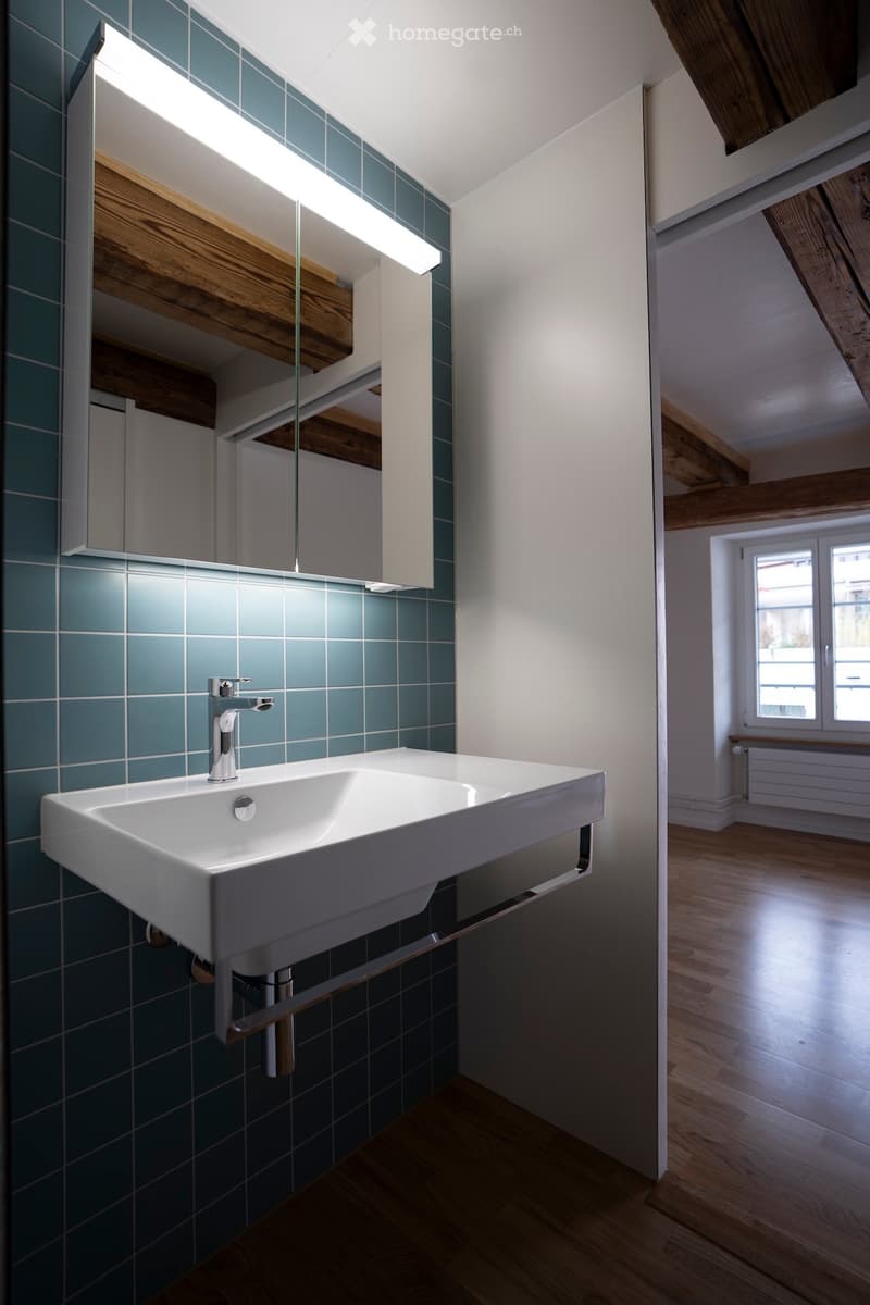Modern bathroom with a white sink, chrome faucet, and a large mirror with built-in lighting. The walls are tiled in a teal color, and the floor is made of light wood.