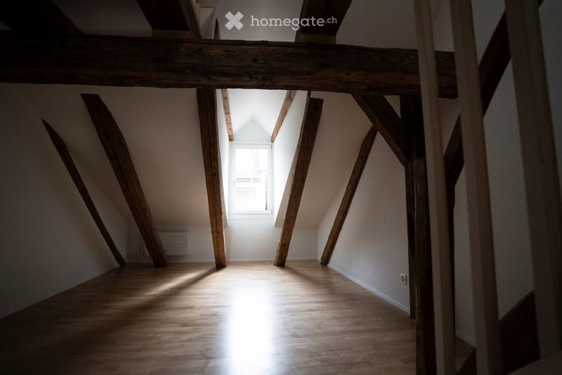 An empty attic space with wooden beams, wooden floor, and a small window letting in natural light.