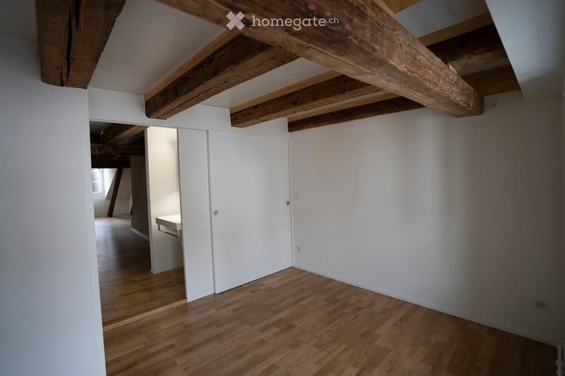 Empty room with exposed wooden beams on the ceiling, white walls, and wooden flooring. A white sliding door leads to another room.