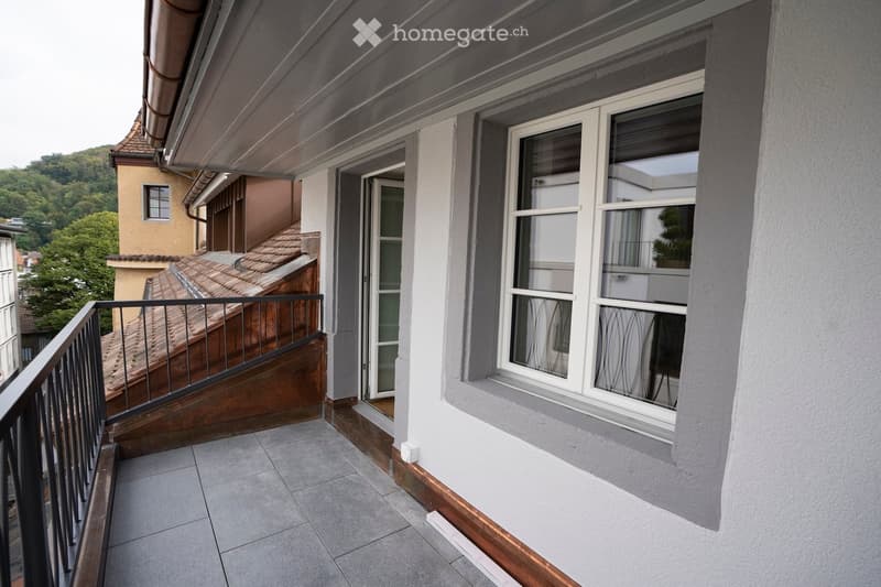 Balcony with tiled floor, metal railing, and access to a wooden staircase leading to a roof area. The building has white walls and large windows.