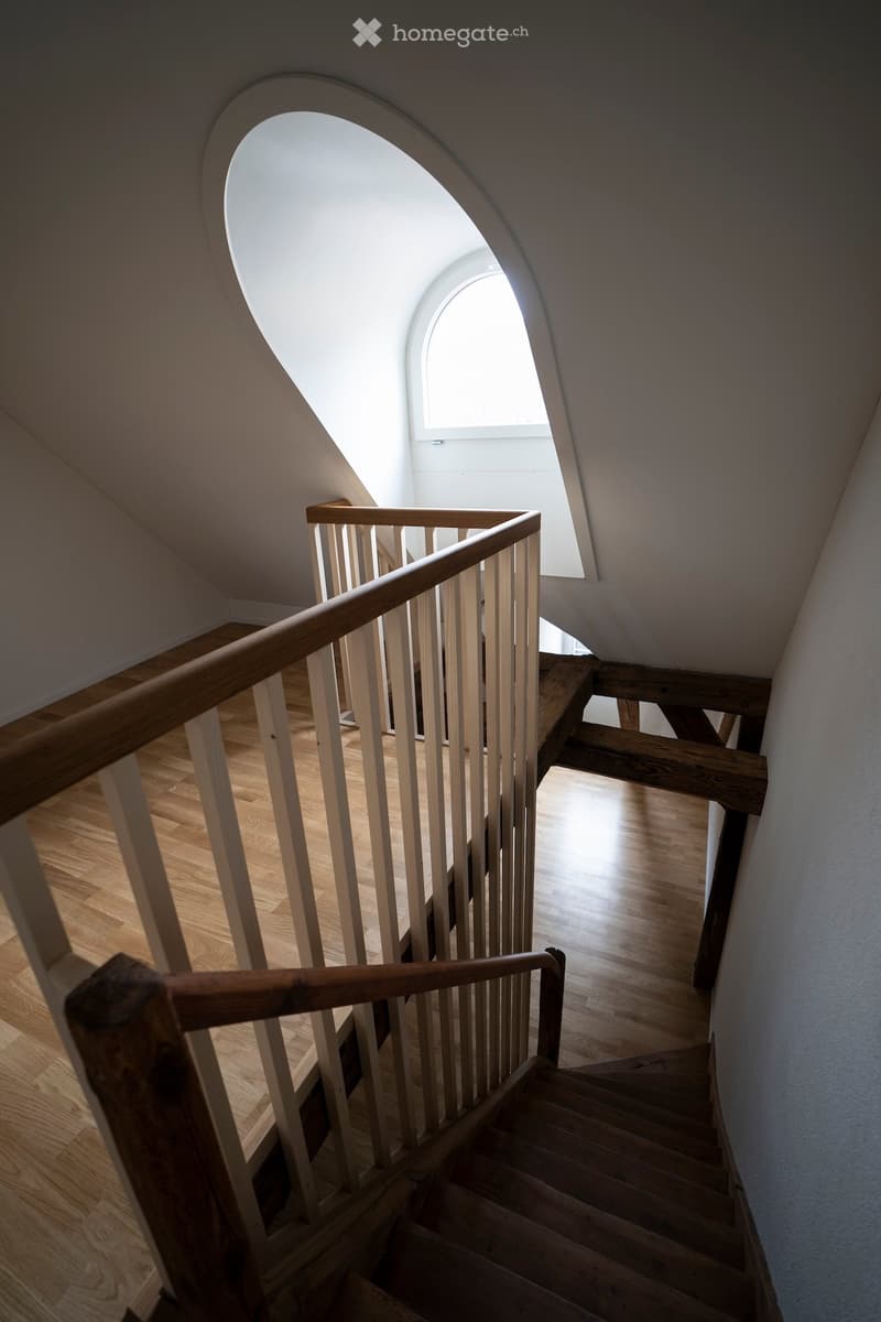 Wooden staircase with a handrail, light-colored wooden steps, and a skylight above, leading to an open area with wooden flooring.