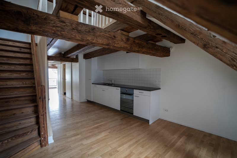 Modern kitchen with white cabinets, black countertop, and stainless steel sink and oven. Exposed wooden beams and staircase visible.
