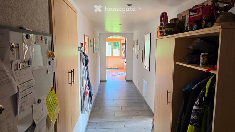 A well-lit hallway with tiled flooring, wooden cabinets, and a coat rack. A doorway leads to another room with a visible rug and window.