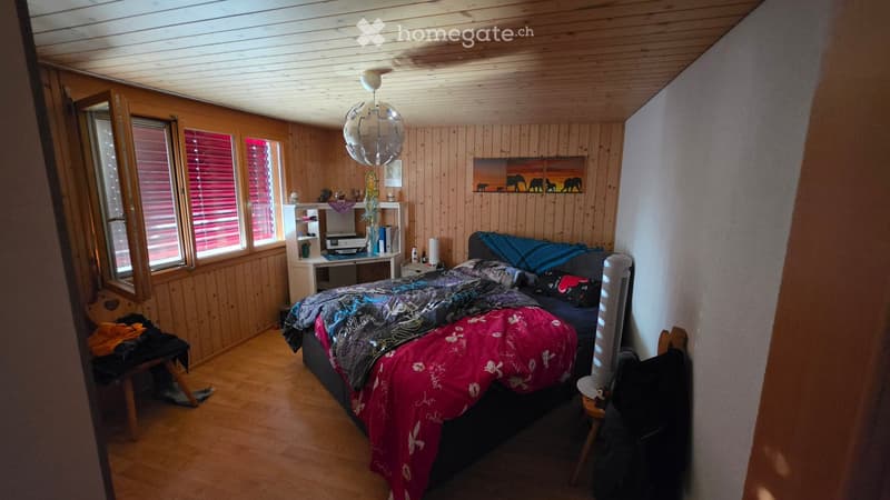 Cozy bedroom with wooden walls and ceiling, a bed with colorful bedding, a nightstand, a fan, a chair, and a shelf with various items.