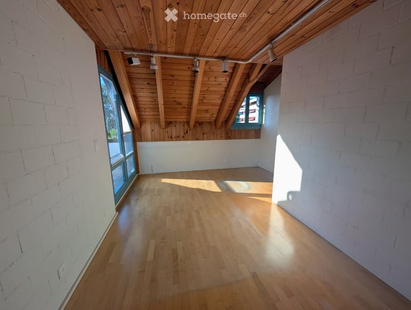 Empty room with wooden flooring, white brick walls, and exposed wooden beams on the ceiling. Natural light streams in through large windows, casting shadows on the floor.