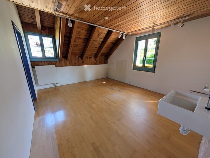 Empty attic room with wooden floors, exposed wooden beams, white walls, and two windows. A white sink is visible in the corner.