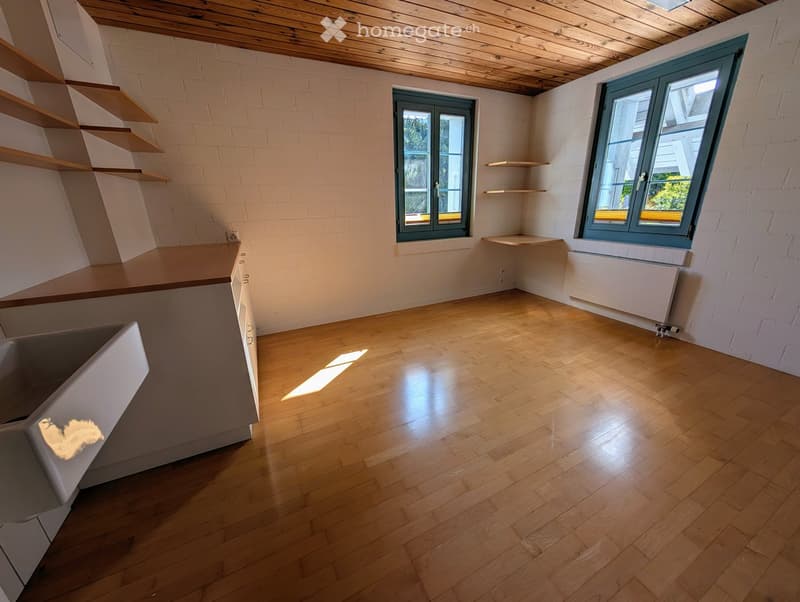 Empty room with wooden flooring, white brick walls, and built-in shelving units. Two windows with green frames allow natural light to enter.