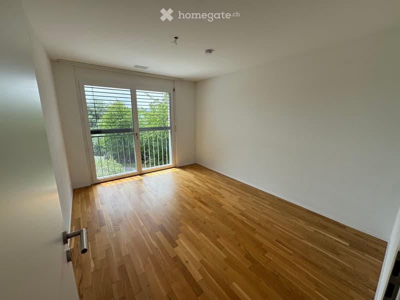 Empty room with light wooden flooring, white walls, and a large sliding glass door leading to a balcony with a metal railing.
