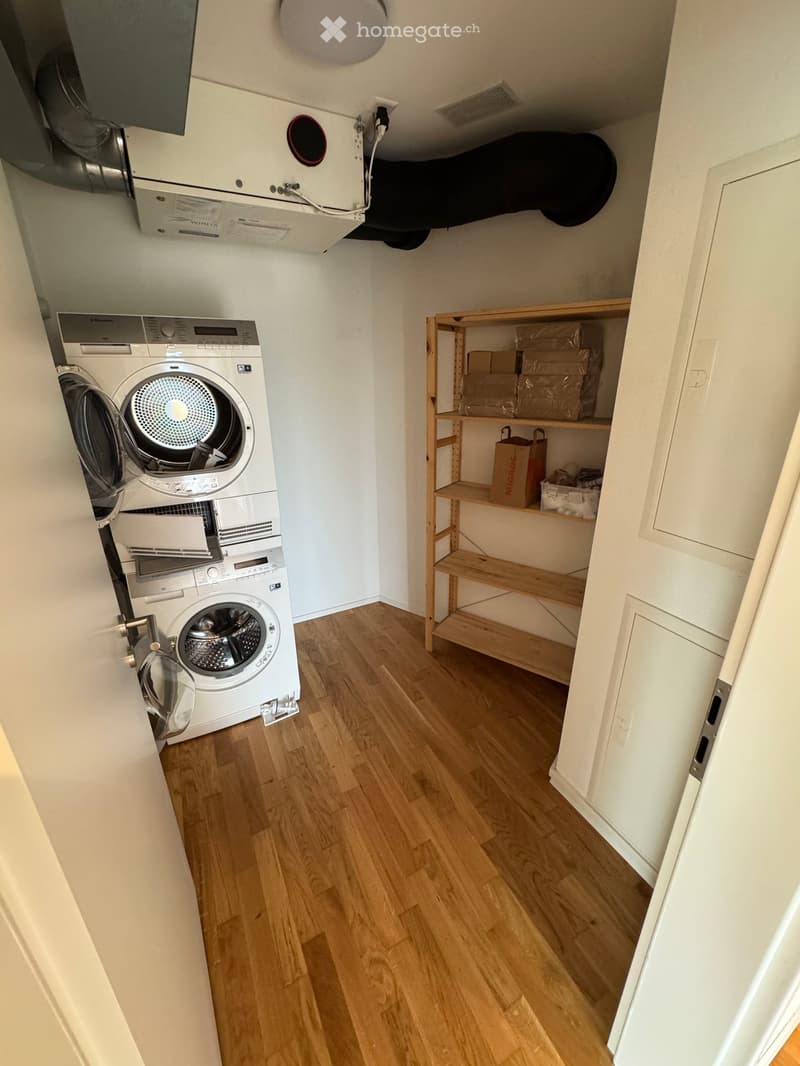 Small room with wooden flooring, white walls, a washing machine and dryer unit, and a wooden shelf with storage boxes and bags.