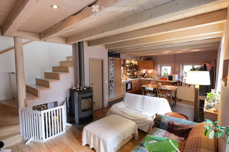 Open-plan living and dining area with exposed wooden beams, a covered sofa, ottoman, dining table with chairs, kitchen area with cabinets and appliances, and a wood-burning stove.