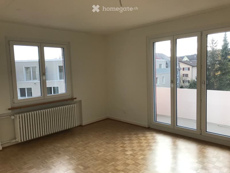 Empty room with wooden flooring, large windows, radiator, and sliding glass doors leading to a balcony.