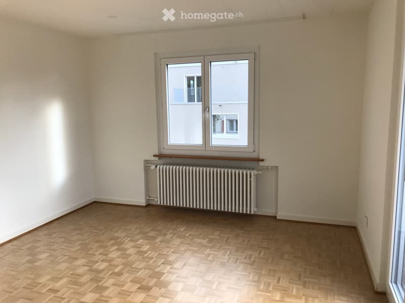 Empty room with wooden flooring, white walls, a radiator, and a large window with a view of a neighboring building.