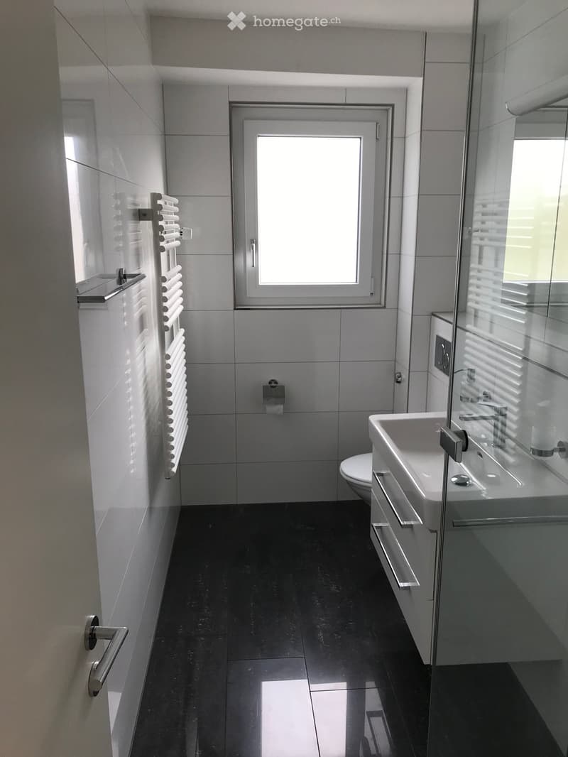 Modern bathroom with black tiled floor, white tiled walls, a sink, toilet, radiator, and a large window with blinds.
