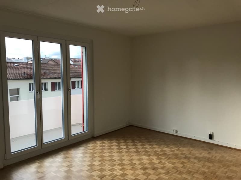 Empty room with wooden parquet flooring and large sliding glass doors leading to a balcony or outdoor area. The walls are plain and there are no furnishings or decorations visible.