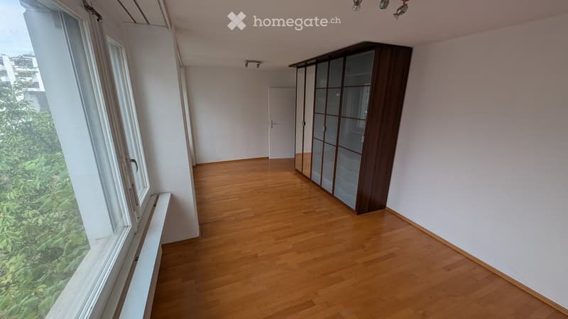Empty room with wooden flooring, white walls, large window with view of greenery, and a tall wardrobe with glass doors.