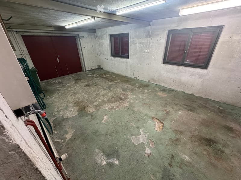 Empty basement space with concrete walls and floor, two windows with blinds, a large red door, and some pipes and hoses visible.