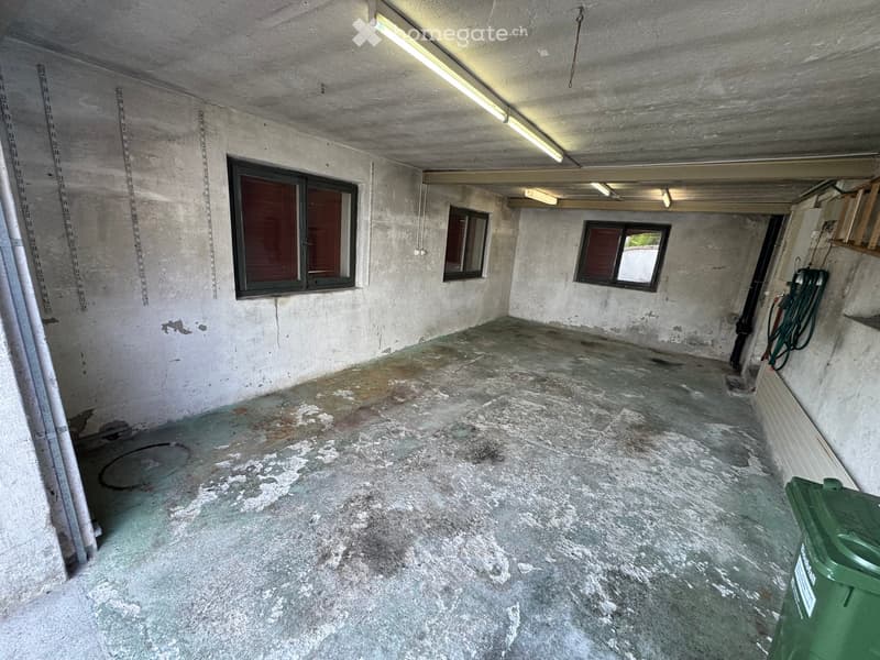 Empty concrete room with three windows, exposed pipes, and a green trash bin in the corner.