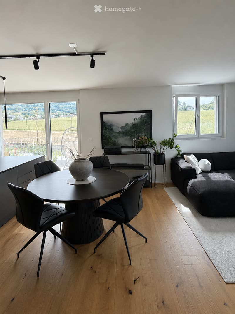 Modern living room with light wooden flooring, a black sofa, a round black coffee table with a vase, and a black dining table with chairs. Large windows offer a scenic view.