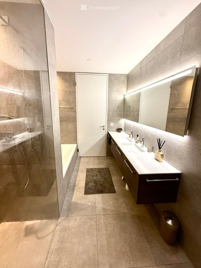 Modern bathroom with a large mirror, double sink, bathtub, and separate shower area. Features include built-in cabinets, sleek fixtures, and a neutral color palette.