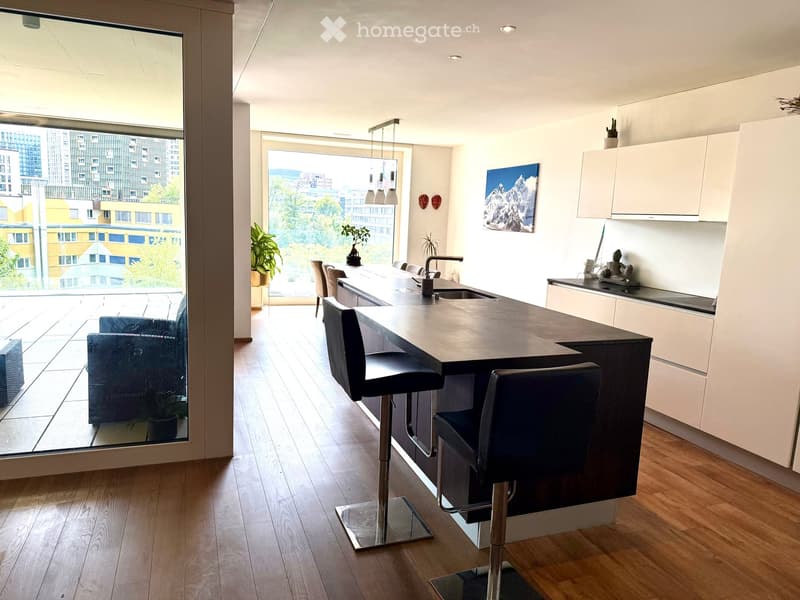 Modern kitchen with a central island, bar stools, a sofa, and a view of the city through large windows.